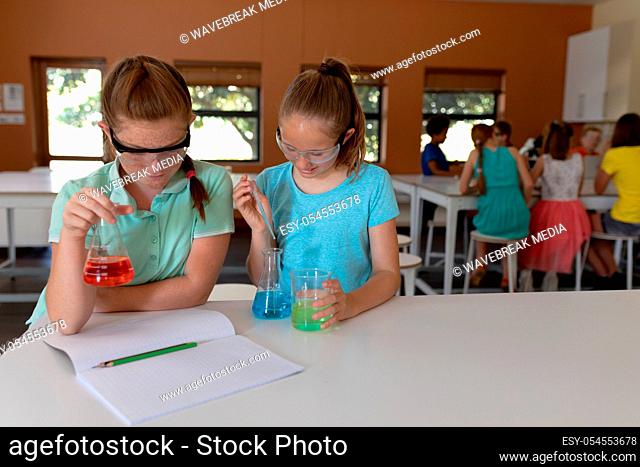 Front view of two Caucasian schoolgirls sitting at a desk wearing safety glasses and using flasks of colourful liquid while doing an experiment during an...