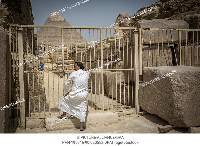 16 July 2019, Egypt, Giza: A Egyptian man stands in front of a closed gate backdropped by the Sphinx and the Pyramid of Khafra at the Giza Pyramids complex