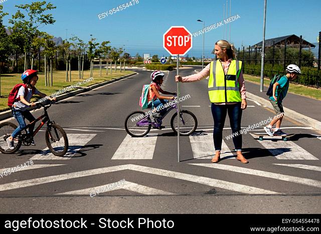 Front view of a blonde Caucasian woman wearing a high visibility vest and holding a stop sign, standing in the road, turning to the side while three...