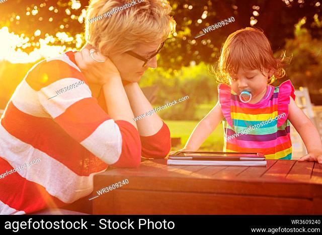 Happy mother and her little daughter enjoying free time using tablet computer while relaxing on holiday home garden during sunny day
