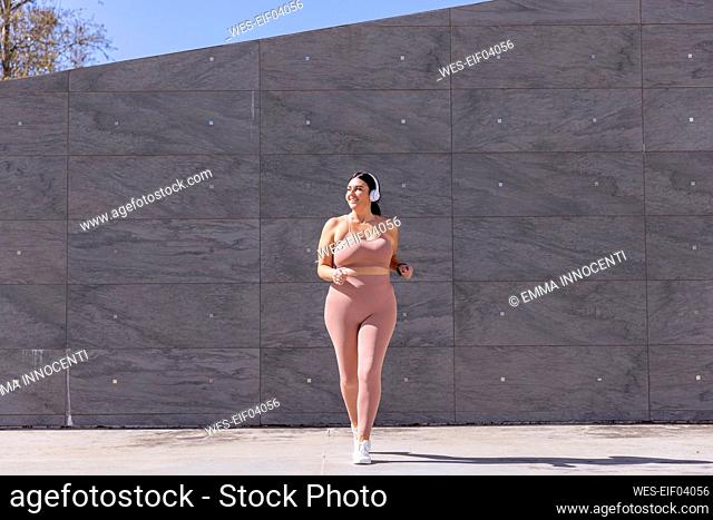 Smiling plus size woman listening music through wireless headphones walking in front of wall