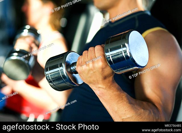 Strong man exercising with dumbbells in a gym, in the background a woman also lifting weights; focus on hand and dumbbell