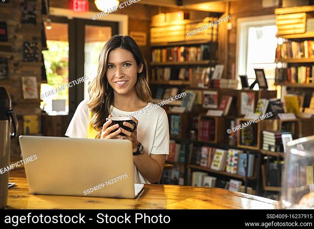 Portrait of young woman holding coffee mug, sitting at counter of coffee shop bookstore with laptop computer