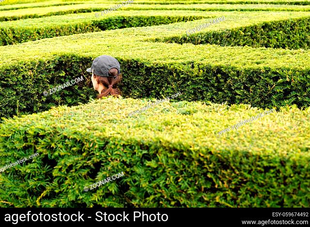 A woman wearing a baseball cap walks around lost in a giant labyrinth made of boxwood hedges