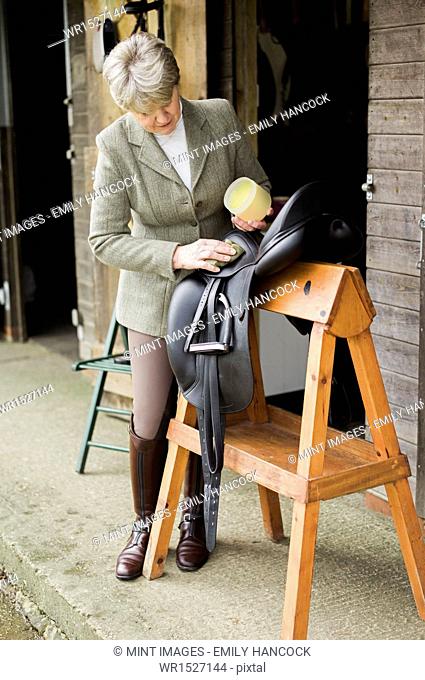 A woman cleaning and preparing tack and saddle, in the courtyard of a riding stable