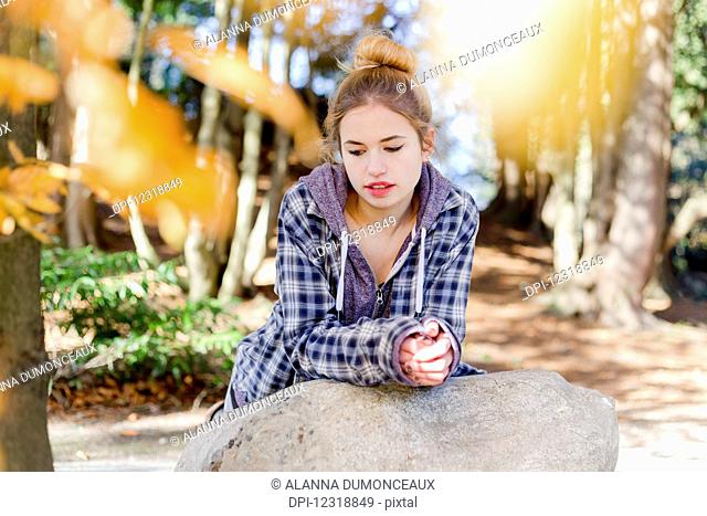 This young teenage girl hangs out alone in a park, sitting on a rock in a thoughtful disengaged position thinking to herself; New Westminster, British Columbia