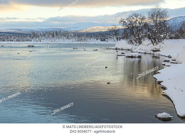 Winter landscape in Tjåmotis with mountains in background, sun shining over the mountains, creek with open water, sky with nice colors reflecting in the water
