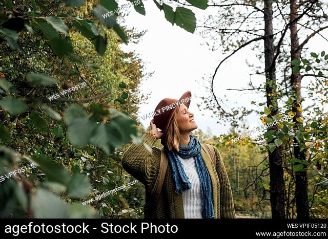 Woman adjusting hat while standing with eyes closed in forest