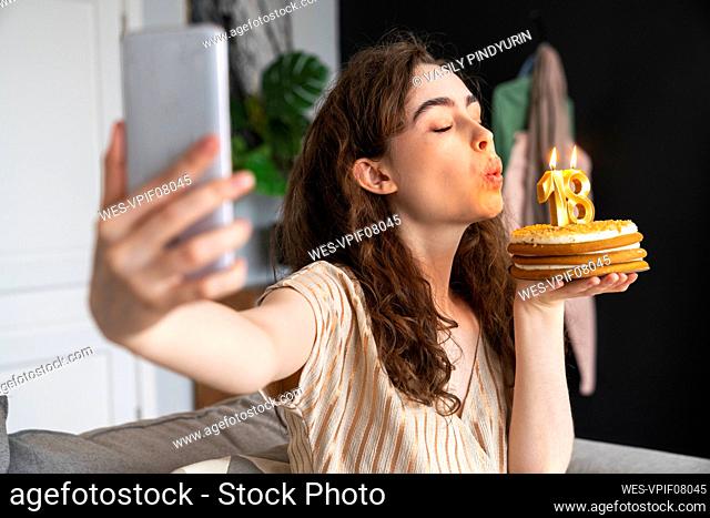Young woman blowing candle on birthday cake and taking selfie at home