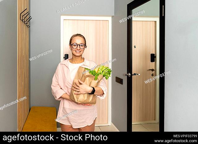 Smiling woman holding vegetables in paper bag at home