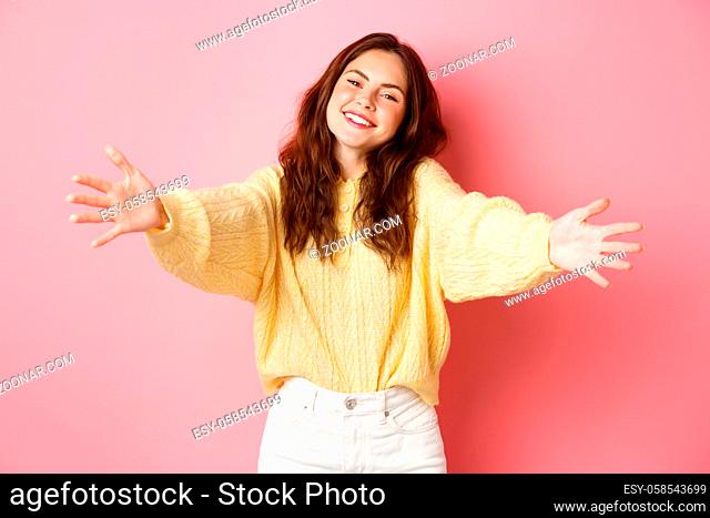 Young beautiful woman reaching hands forward to hug you, smiling friendly, inviting for free cuddles, standing against pink background