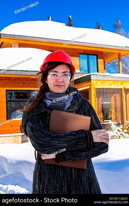 vertical portrait of a construction architect woman wearing a red hard hat and holding clipboard. she standing on the front of snow covered building