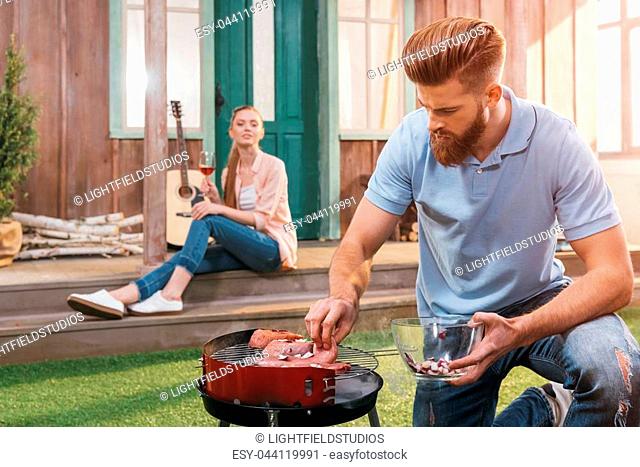 bearded man roasting meat on barbecue grill with woman with wine behind