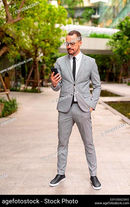 Portrait of handsome Hispanic bald bearded businessman wearing suit in the city with nature outdoors