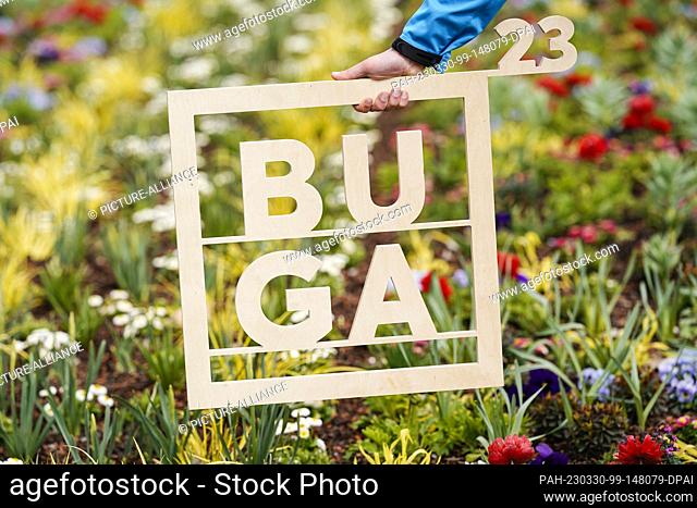 ILLUSTRATION - 30 March 2023, Baden-Württemberg, Mannheim: A hand holds a wooden frame with the inscription ""BUGA 23"" in front of a flower bed on the grounds...