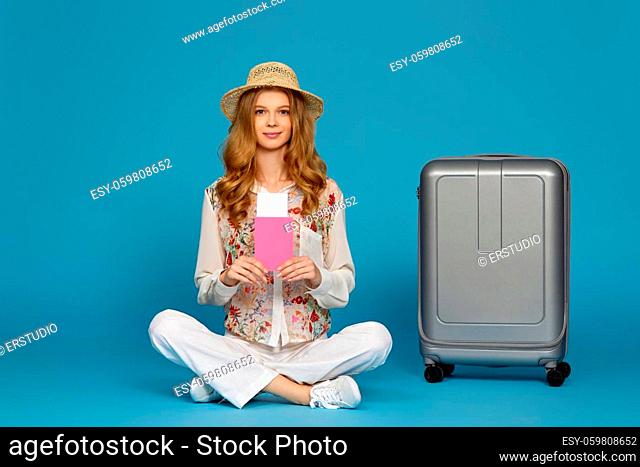 Happy blonde curly woman in hat sitting near suitcase on blue background. copy space