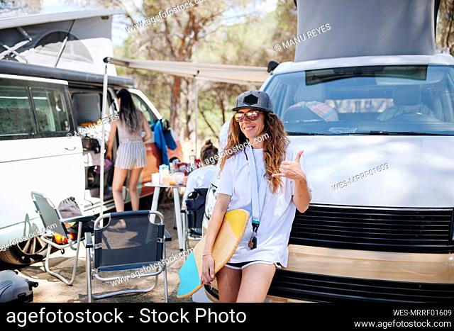 Smiling woman with skateboard gesturing while leaning on camper van