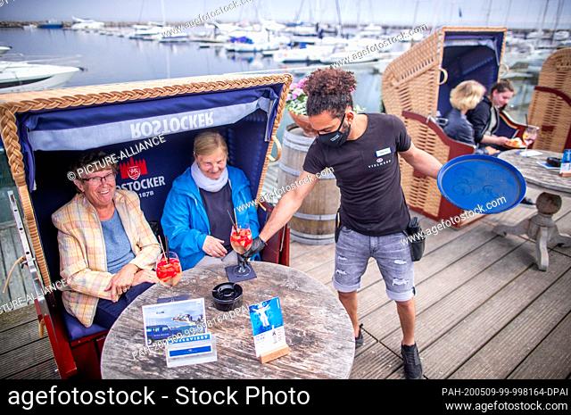 09 May 2020, Mecklenburg-Western Pomerania, Kühlungsborn: A waiter with a face mask serves drinks to two women in a wicker beach chair on the terrace of the...