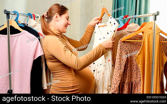 Pregnant smiling woman choosing and trying dresses hanging on clothes rack in her wardrobe at bedroom