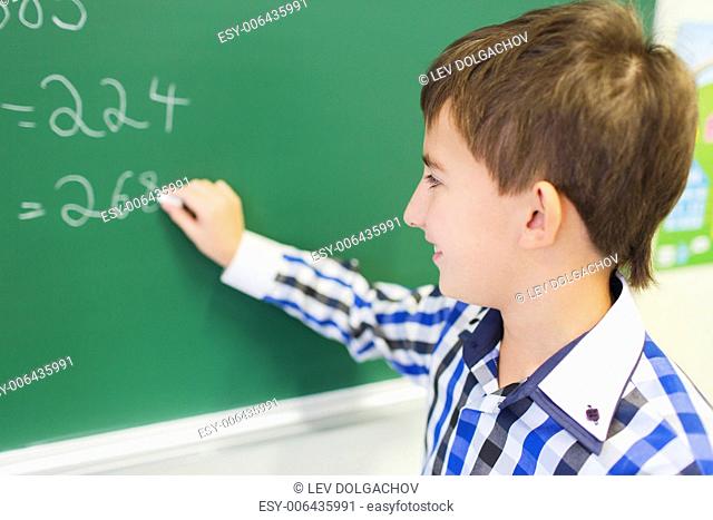 education, elementary school, learning, math and people concept - little smiling schoolboy writing numbers on green chalk board in classroom