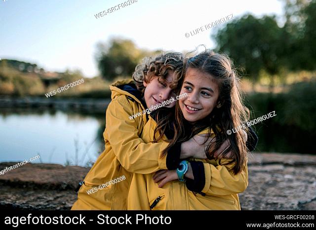 Boy hugging smiling sister wearing yellow raincoats at riverbank