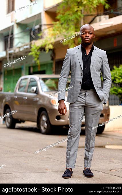 Portrait of young handsome bald African businessman wearing suit in the streets outdoors