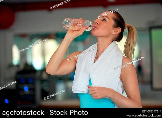Middle-aged woman athlete drinking bottled water for hydration after working out in a gym in a health and fitness concept