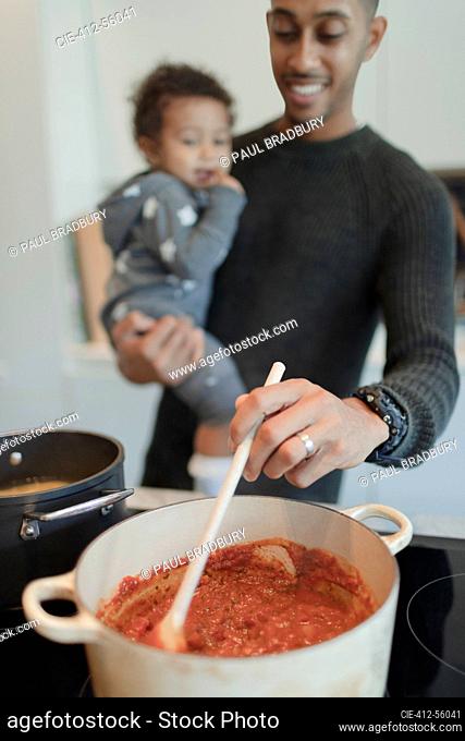 Father holding baby daughter and cooking spaghetti at stove