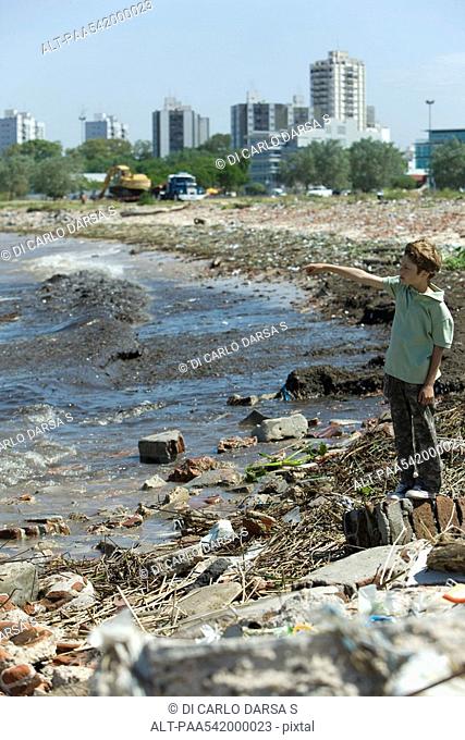 Boy standing on polluted shore, looking at view, pointing