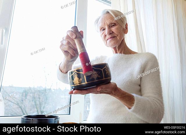 Smiling senior woman with singing bowl standing by window at home