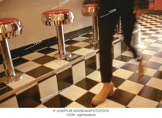 Woman in black pants and red shoes walking on a black and white tiled floor in a diner