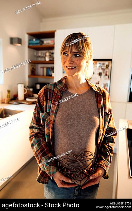 Smiling expectant woman holding heart shape model standing at home