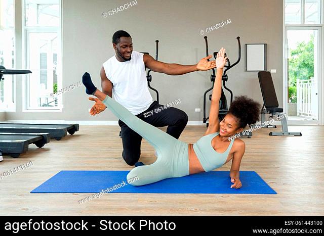 Short curly black hair coach with moustache and beard teach young woman in sportswear how to do side plank crunch. Cardio machines are on the background at the...