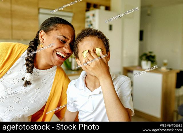 Happy mother embracing son covering eyes with cucumbers at home
