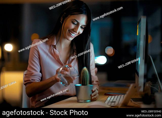 Smiling young woman sitting at desk in office watering cactus