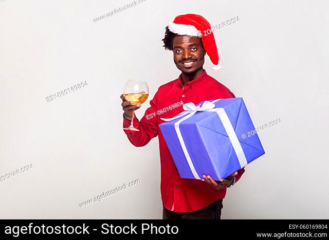 Happiness afro man in red cap holding champagne glass and gift box, looking at camera and toothy smiling. Studio shot. Gray background