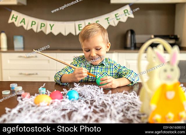 Joyous busy cute little boy seated at the table coloring eggs with a wooden brush