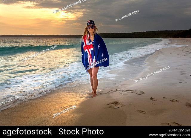 Woman with the Australian flag wraped around, wears a hat and sunglasses also with the Australian flag, as she walks along the wet sand of the beach in early...