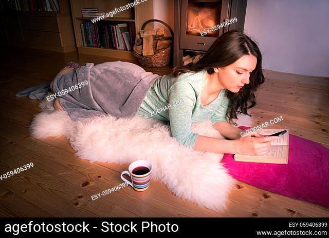 Young brunette girl relaxing in front of the fireplace, lying on a white fur, on the floor, while using a smart phone, having an open book and a cup of tee near...
