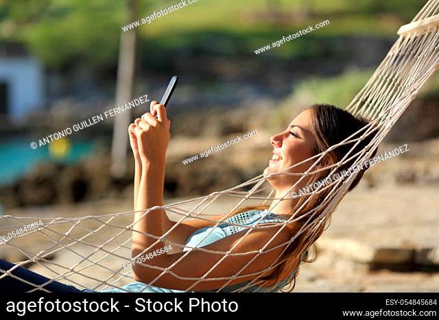 Side view portrait of a happy woman checking smart phone relaxing lying on a hammock on the beach