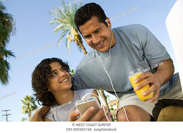 Boy 13-15 holding portable music player father listening with earphones and holding glass of juice