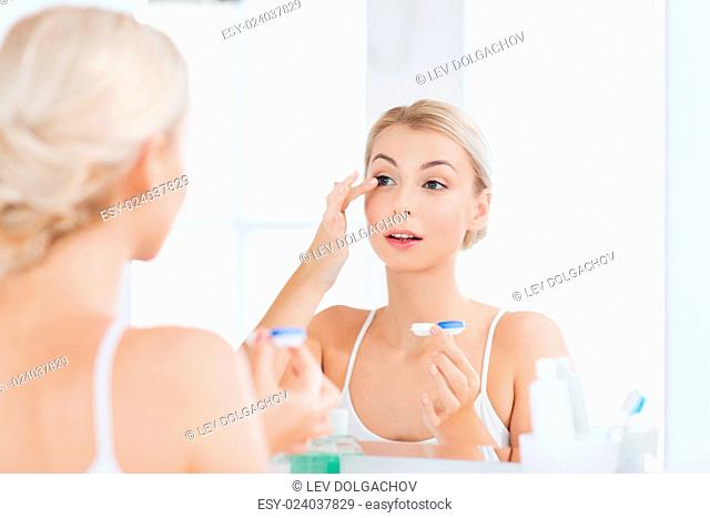 beauty, vision, eyesight, ophthalmology and people concept - young woman putting on contact lenses at mirror in home bathroom