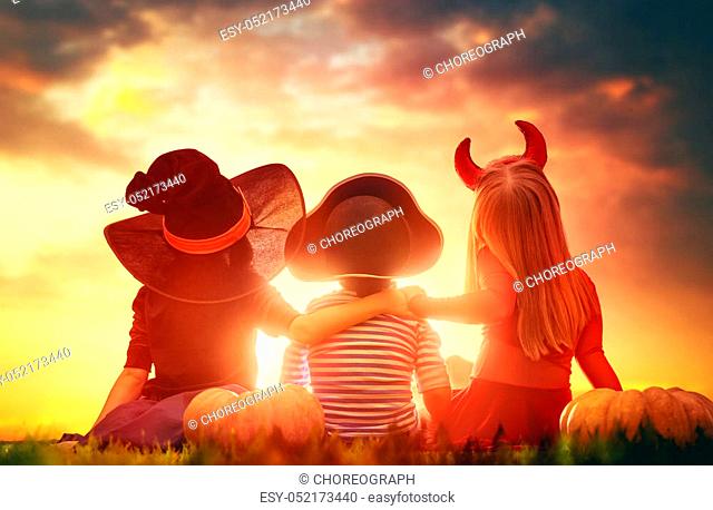 Happy brother and two sisters on Halloween. Funny kids in carnival costumes outdoors. Cheerful children and pumpkins on sunset background