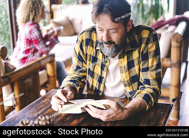 Adult man reading a book at home sitting on the wooden table with wife woman in background. Happy caucasian couple of people enjoying leisure time indoor...