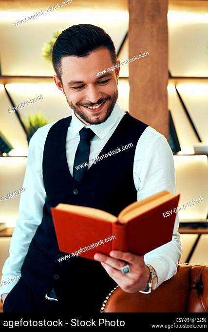 Handsome young man in elegant suit reading a book in modern luxury interior. Hobby concept