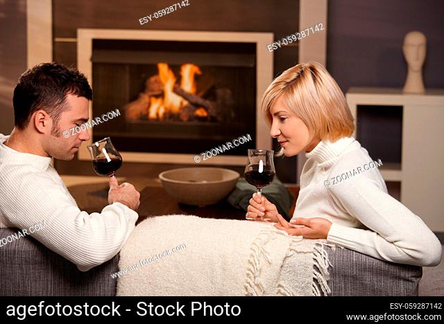Young romantic couple sitting on sofa in front of fireplace at home, drinking red wine