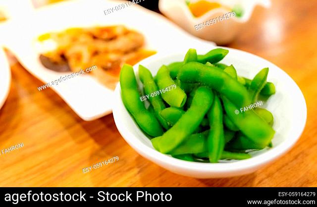 Edamame or green soybeans blanched in white bowl on wooden table at Japanese restaurant. Pods of green soy beans on blurred Japanese food