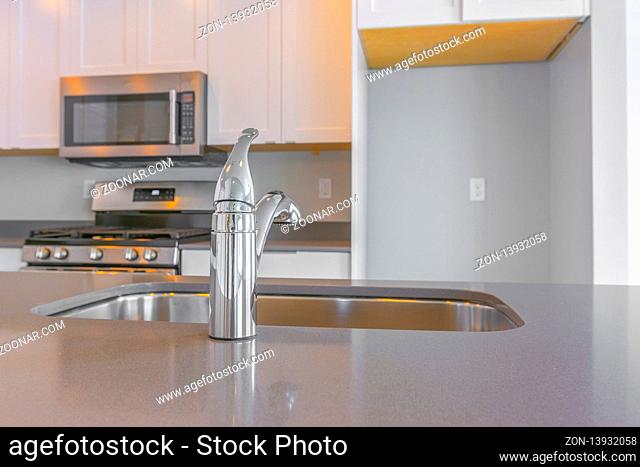 Modern kitchen interior with close up on the shiny faucet and sink. A cooking range, microwave, and empty refrigerator alcove can be seen in the background