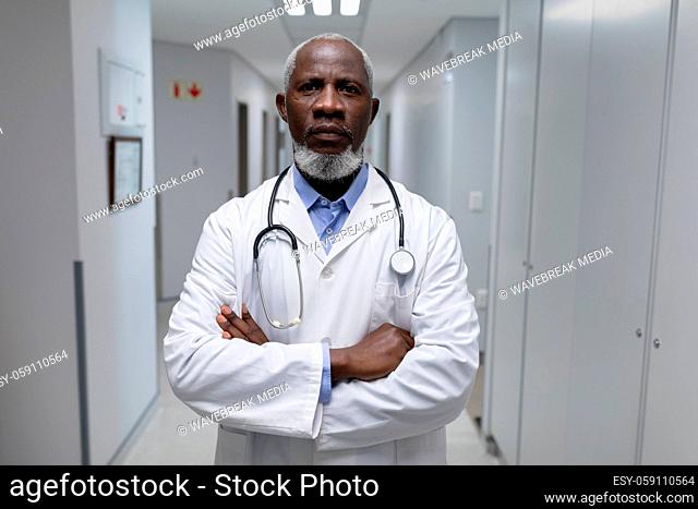Portrait of senior male doctor with stethoscope wearing lab coat, arms crossed in hospital corridor