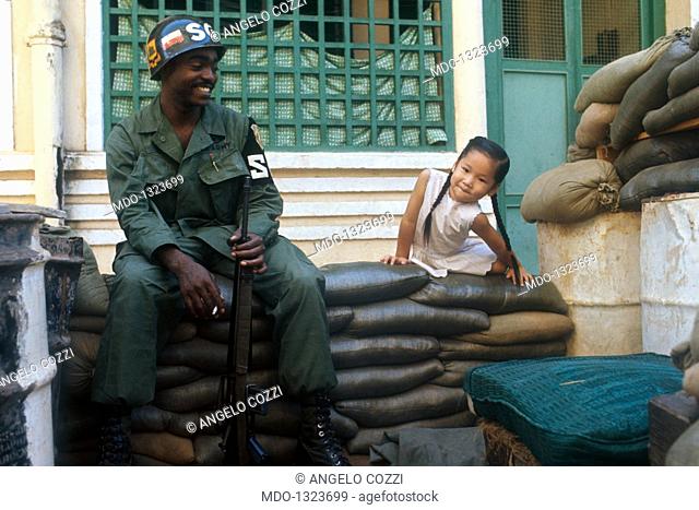 A smiling US soldier next to a young Vietnamese girl. A soldier from the SG division of the US army amused as he watches a little Vietnamese girl as she climbs...